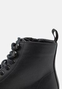 Black leather ankle boot with round toe, textured surface, black metal eyelets, and gray laces. Features a fabric pull tab at the rear.