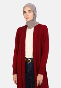 Burgundy long cardigan with open front, ribbed texture, worn over a white top. Accompanied by a taupe hijab and dark denim jeans.