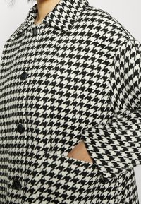 Płaszcz w czarno-białą kraciastą kratkę houndstooth z kołnierzykiem, trzema guzikami i kieszeniami po bokach, prezentujący teksturowany materiał.