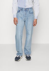 Person som bär ljusblå raka jeans, en blå och vit randig skjorta och svartvita sneakers, stående på ett enfärgat vitt golv.