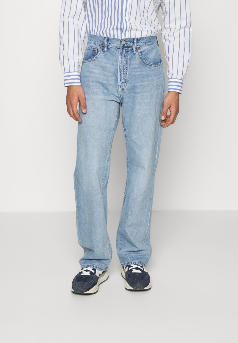 Person som bär ljusblå raka jeans, en blå och vit randig skjorta och svartvita sneakers, stående på ett enfärgat vitt golv.