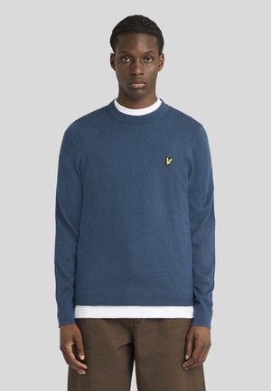 Jeune homme portant un pull bleu à manches longues avec un logo d'oiseau jaune, une chemise blanche en dessous et un pantalon marron, debout devant un fond gris.