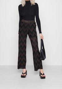 Kobieta ubrana w czarną bluzkę z długim rękawem, szerokie czarne spodnie ze złotym wzorem w zygzak, trzyma czarną torebkę, stoi w sandałach na platformie.