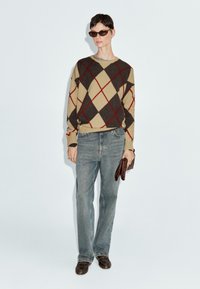 Pull en argyle beige avec des diamants rouges et marron, associé à un jean droit bleu clair et des chaussures en cuir marron. Tenant une pochette marron.