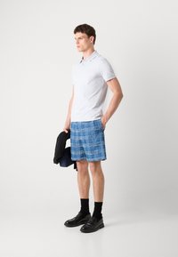 Λευκό πόλο μπλουζάκι, μπλε καρό σορτς με φθαρμένο τελείωμα, μαύρες loafers και μαύρες κάλτσες. Το μοντέλο κρατά μια μαύρη ζακέτα. Μινιμαλιστικός σχεδιασμός.