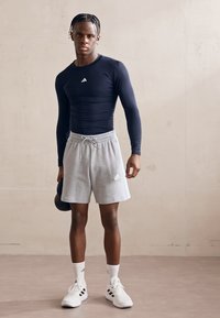 Juoda ilgomis rankovėmis sportinė maikutė, pilkos sportinės šortai, balti saitai ir sportbačiai. Laiko kettlebell'ą. Paprastas dizainas, logotipas ant drabužių.