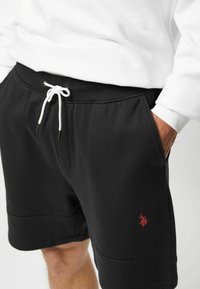 Zwarte katoenen sportshorts met een elastische tailleband, witte trekknoop, zijzakken en een klein rood logo op het linkerpand.