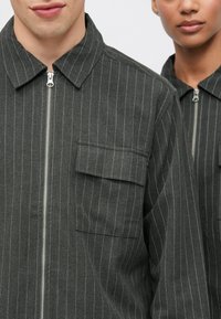 Ciemnoszare, cienko prążkowane marynarki na zamek błyskawiczny, z kołnierzem i naszywaną kieszenią, wykonane z teksturowanej tkaniny. Zbliżenie na dwie osoby.