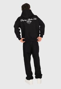 Personne portant un sweat à capuche noir et un pantalon, avec des baskets blanches, de dos, montrant le texte du sweat "Dropsize Studios Ltd. Le succès est un jeu de rue."