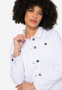 Chaqueta de mezclilla blanca con un diseño ajustado, botones negros con un patrón de estrellas y sutiles detalles desgastados. Presenta bolsillos de solapa y puños enrollados.