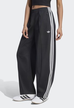 Persona che indossa pantaloni della tuta Adidas neri a gamba larga con strisce bianche e scarpe Adidas bianche, in piedi davanti a uno sfondo semplice.
