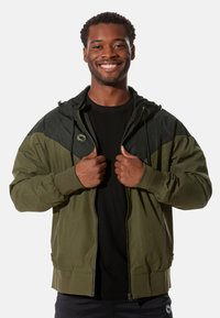 Olivgrüne Kapuzenjacke mit schwarzem Oberteil, ausgestattet mit einem Reißverschluss, elastischen Bündchen und einer strukturierten Stoffoberfläche.