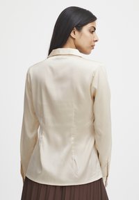 Cremfarbendes Satin-Hemd mit langen Ärmeln und Kragen, taillierter Schnitt, rückwärtige Abnäher und knöpfbare Manschetten. Glatte Textur und dezenter Glanz.