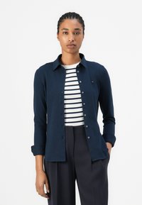 Camicia button-up blu con maniche lunghe, realizzata in tessuto liscio. Presenta un colletto classico e un logo del marchio discreto sul petto.