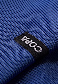 Blauwe ribstof met een gestructureerd oppervlak. Een zwart label met het witte woord "Copa" is in de stof genaaid en is schuin geplaatst.