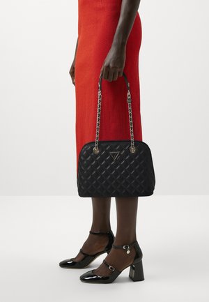 Sac à main en cuir matelassé noir avec quincaillerie dorée, doté de chaînes. Associé à des talons en cuir verni noir et à une robe rouge texturée.