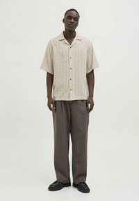 Srajca s kratkimi rokavi z bež vzorcem v obliki diamantov in lesenimi gumbi, kombinirana z ohlapnimi sivimi hlačami in črnim obutjem.