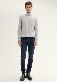 Svetlosivý rolákový sveter z jemnej látky, skombinovaný s tmavomodrými slim-fit džínsami a čiernymi členkovými čižmami s hladkým povrchom.