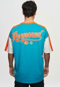 Turkos- och orangefärgad kortärmad t-shirt med en stor "Recovered" grafisk design på baksidan, med kontrasterande ärmar och textil med struktur.