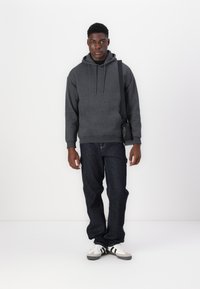 Hanorac gri, blugi închis și adidași albi cu dungi negre. Hanoracul are o buzunar frontal și un glugă cu șnur. Design simplu.