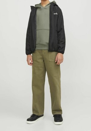 Personne portant une veste noire zippée, un sweat à capuche vert olive, un pantalon assorti vert olive et des baskets noires avec des semelles blanches.