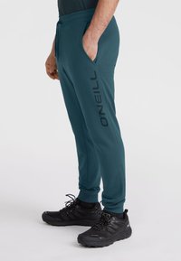 Turkusowe dresowe spodnie z bocznymi kieszeniami, elastycznym pasem i wyraźnym czarnym brandingiem "O'Neill" na lewej nogawce. Noszone z czarnymi butami sportowymi.