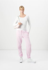 Woman standing with hand in pink sweatpants pocket, wearing white long-sleeve top, white sneakers, and carrying a light pink shoulder bag.