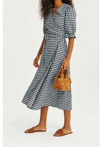 Femme portant une robe midi à carreaux noir et blanc avec des manches bouffantes, tenant un petit sac tressé beige et portant des sandales plates marron.