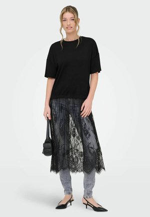 Ung kvinne iført en løs svart topp, gjennomsiktig svart blonderkjørt over grå jeans, svarte slingback hæler, og holder en liten svart håndveske.