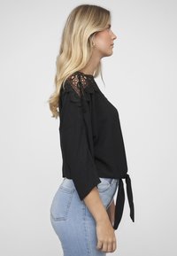 Svart topp med heklet blonderdetaljer på skuldrene, avslappet passform, trekvart ermer og knyttet midje. Kombinert med lyseblå jeans.