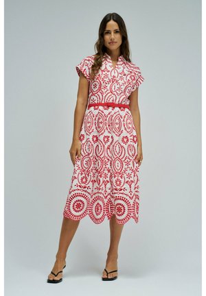 Mujer de pie con un vestido midi blanco con estampados florales rojos y un cinturón rojo, combinado con sandalias negras de tacón, sobre un fondo liso.