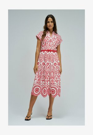 Femme debout portant une robe midi blanche à motifs floraux rouges avec une ceinture rouge, associée à des sandales à talons noires sur un fond uni.