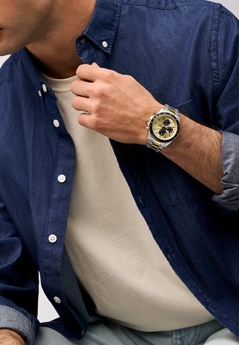 Srebrny i złoty zegarek chronograf z trzema czarnymi tarczami na stalowej bransolecie noszony pod niebieską koszulą z guzikami. Swobodny wygląd.