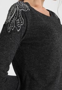 Pull gris avec un tissu texturé orné de sequins sur les épaules, présentant un col arrondi et une coupe ajustée.