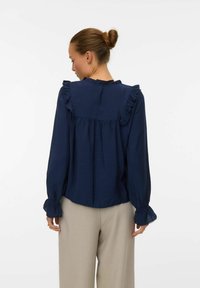 Blusa blu navy a maniche lunghe con dettagli arricciati sulle spalle e sui polsini, caratterizzata da un design a pieghe sul retro in tessuto leggero.