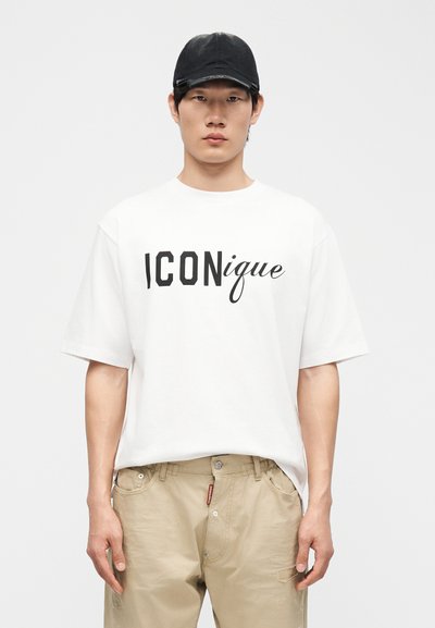 Νεαρός άνδρας που φοράει μαύρο καπέλο, λευκό μπλουζάκι με το κείμενο «ICONique» και μπεζ παντελόνι, στέκεται μπροστά σε απλό φωτεινό φόντο.