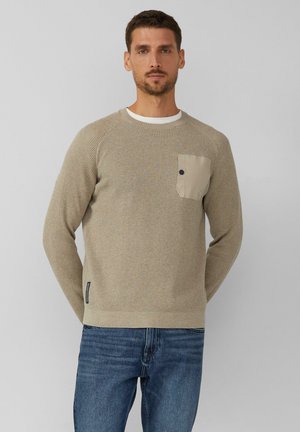 Beige strukturierter Pullover mit Rundhalsausschnitt und einer kontrastierenden hellen Tasche. Verfügt über gerippte Bündchen und Saum. Kombiniert mit blauen Jeans.