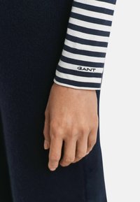 Hand rustend aan de zijde, gekleed in een marineblauwe en witte gestreepte mouw met het "GANT"-logo, gecombineerd met donkere, gestructureerde stofbroek of rok.