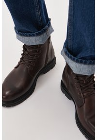 Botas de piel marrón hasta el tobillo con un acabado texturizado, cierre con cordones y suela de goma negra. Jeans de mezclilla azul enrollados visibles por encima.