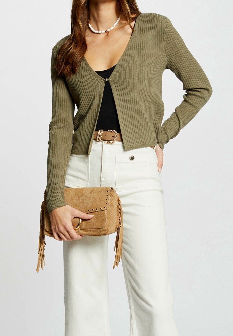 Femme portant un cardigan côtelé vert olive sur un haut noir, pantalon blanc avec une ceinture marron, tenant une pochette en daim beige à franges.