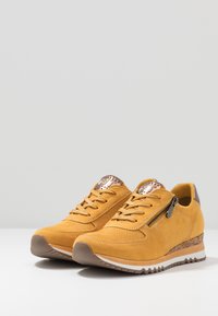 Las zapatillas de ante amarillas cuentan con un diseño perforado, cremallera lateral y acentos metálicos. La suela tiene una textura con una cuña marrón contrastante.