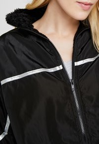 Chaqueta negra con cremallera y un suave y esponjoso cuello, con detalles plateados en los hombros. Tela suave con un acabado semibrillante.