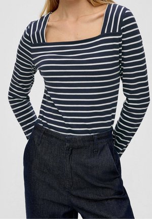 Persoon die een marineblauw-wit gestreept shirt met lange mouwen en een vierkante halslijn draagt, samen met donkerblauwe high-waisted spijkerbroek, handen in de zakken.