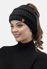 Zwarte gebreide beanie met ribbels en gestikte details. Bevat een bruine label. Draagt met het haar in een paardenstaart gestyled.