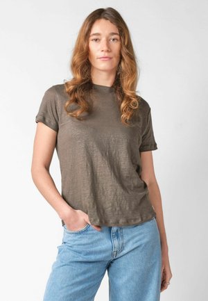 Kobieta z długimi falowanymi włosami stoi z jedną ręką w kieszeni, ubrana w brązową koszulę z krótkim rękawem i jasnoniebieskie dżinsy na białym tle.