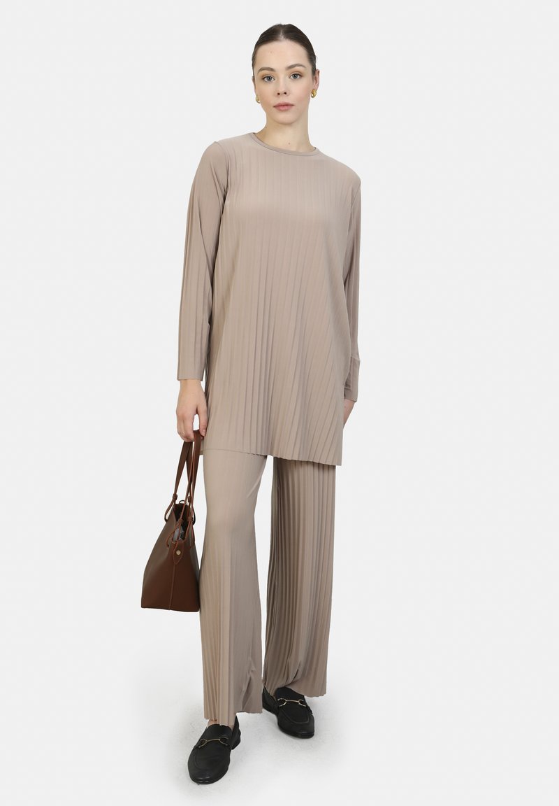 Beige, ribattu, pitkähihaine tuniikki ja leveälahkeiset housut. Malli pitää kädessään ruskeaa käsilaukkua ja käyttää mustia loafer-kenkiä. Suora pinta, moderni muotoilu.