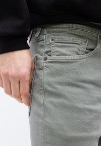 Χέρι που ακουμπά κοντά στην μπροστινή τσέπη ενός ανοιχτόχρ ωμου γκρι τζιν, που φοριέται με μαύρο μακρυμάνικο τοπ.