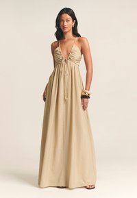 Largo vestido maxi beige con un profundo escote en V, gather frontal y lazo ajustable. Llevado con pulseras negras y doradas y sandalias negras de punta abierta.