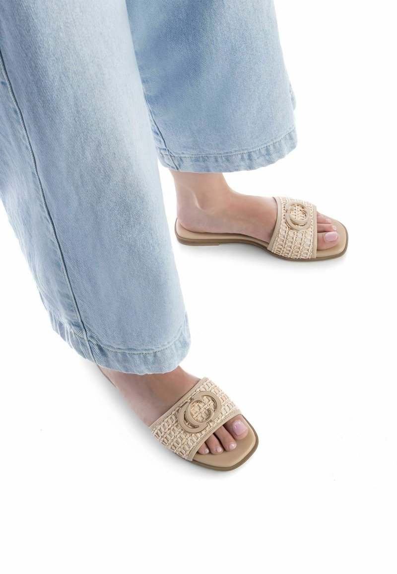 Sandalias deslizantes beige con textura tejida y un acento de logo circular. Suela plana y suave con diseño de punta abierta, combinadas con pantalones anchos de mezclilla claros.