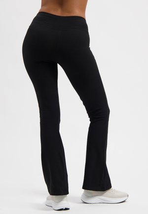 Schwarze, dehnbare Schlaghosen mit hohem Bund, weichem Material und glatten Nähten, getragen mit hellen Sportschuhen.